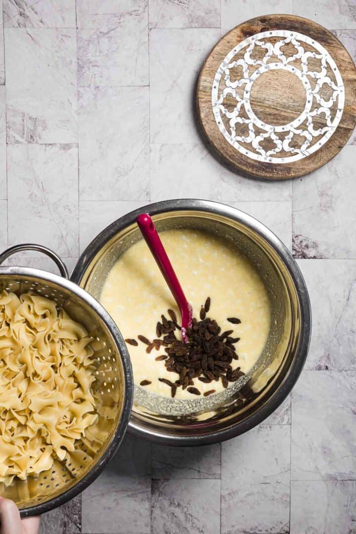 A bowl of cooked egg noodles is added to a large mixing bowl containing a creamy yellow mixture with raisins, crafting a delicious sweet noodle kugel. A stirrer rests in the bowl, all set on a gray tiled surface with a beautifully patterned round wooden trivet nearby.