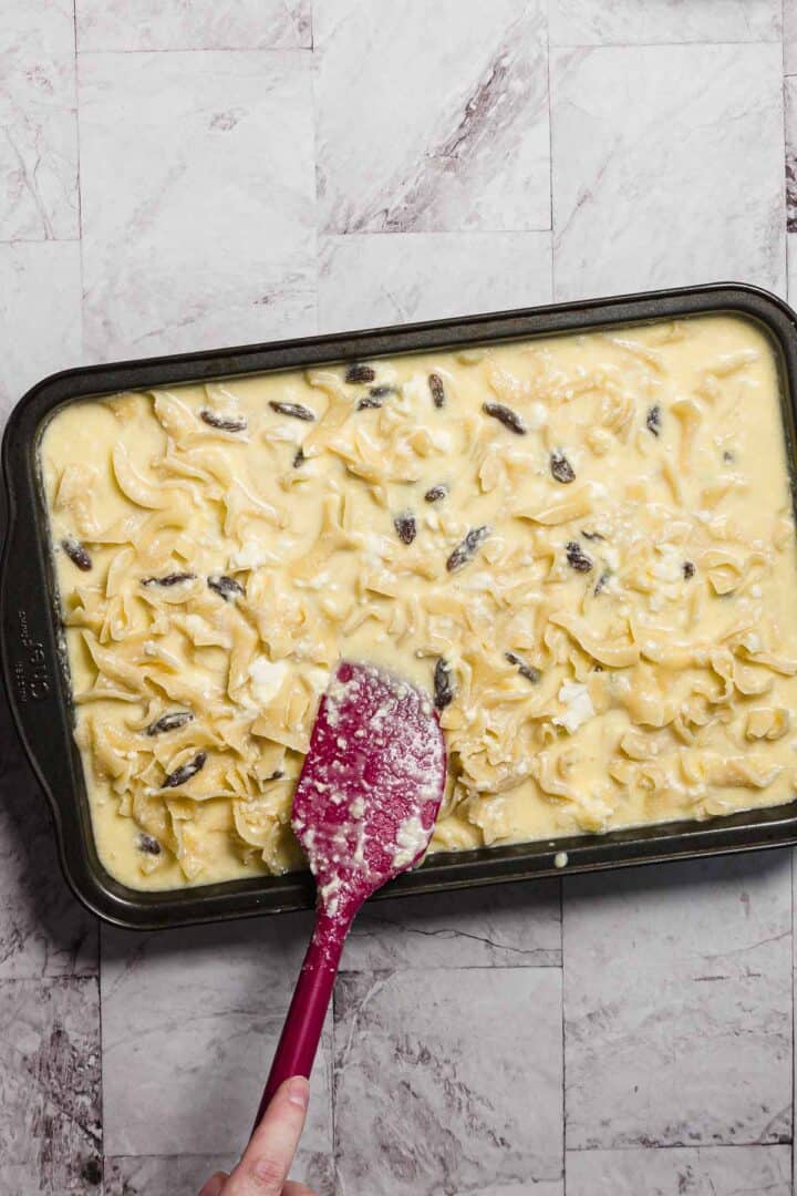 A hand holding a red spatula spreads a creamy sweet noodle kugel mixture with mushrooms in a rectangular baking dish on a marbled countertop.