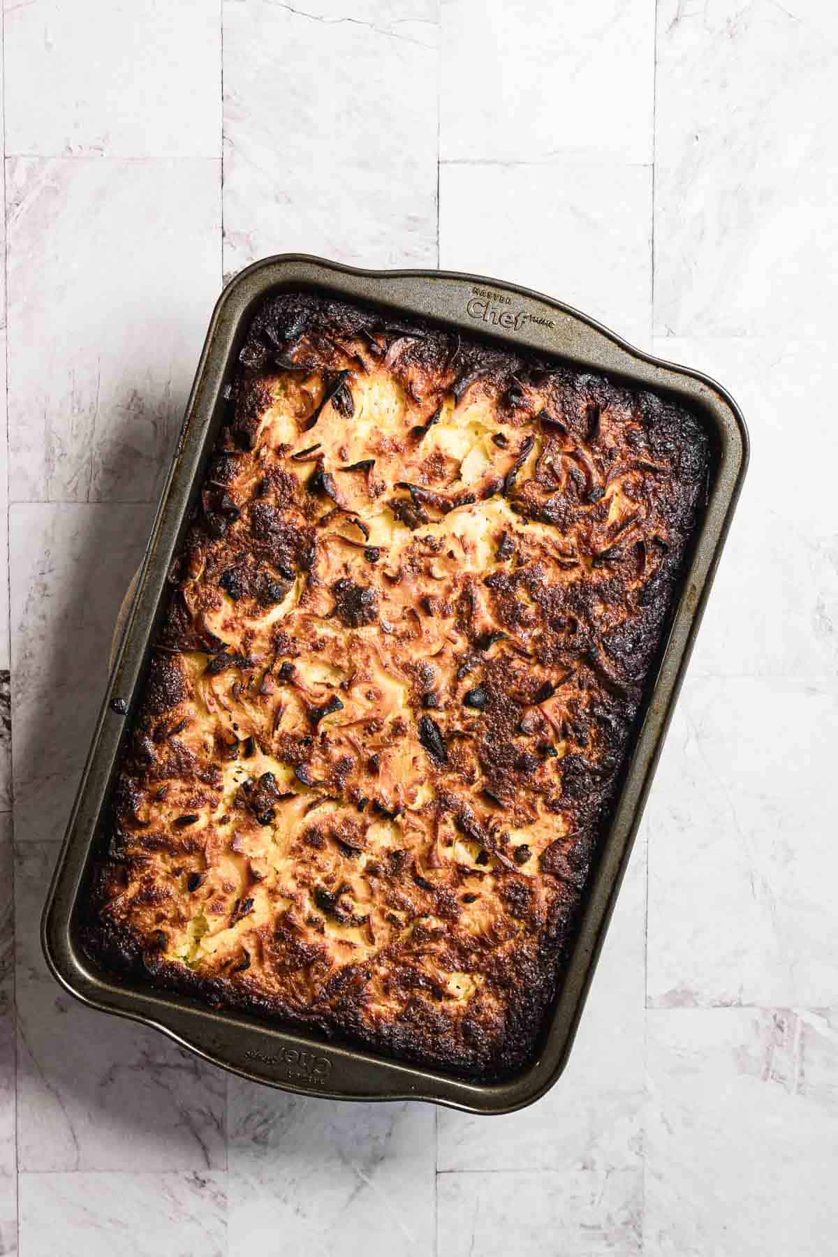 A rectangular baking dish filled with a sweet noodle kugel, its browned, uneven top hinting at a delightful combination of textures, rests on a light-colored tile surface.