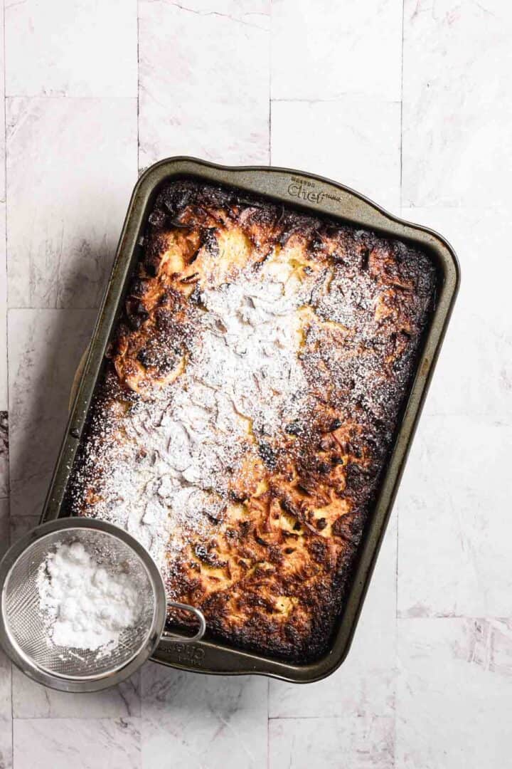 A sweet noodle kugel dessert in a rectangular metal pan sits on a light-colored surface. The golden-brown delight is dusted with powdered sugar, adding a touch of sweetness. Beside the pan, a small sieve with more powdered sugar awaits for an extra sprinkle of indulgence.