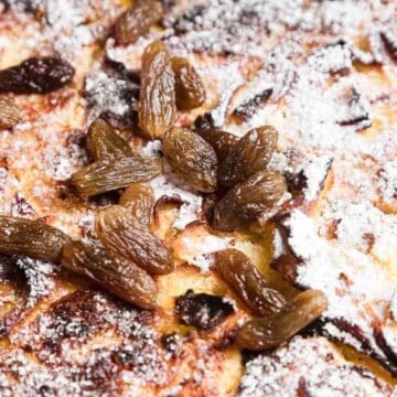 A close-up of a dessert featuring golden-brown flaky pastry dusted with powdered sugar. Topped with a generous amount of raisins scattered across the surface. The texture appears crispy and rich.