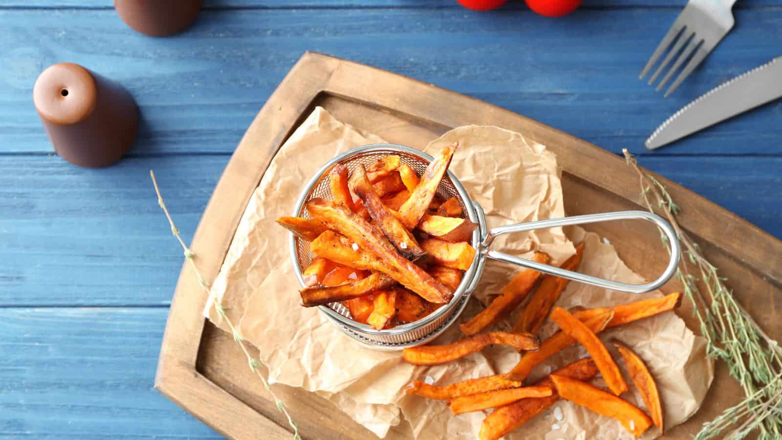 A metal basket filled with sweet potato fries is placed on parchment paper on a wooden board. Some fries are scattered beside the basket. Nearby are sprigs of herbs, a fork, a knife, and salt and pepper shakers on a blue wooden table.