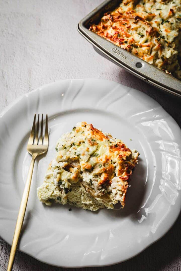 A slice of cheesy casserole with a golden-brown top sits on a white plate beside a gold fork. The creamy, gluten-free dish reveals herbs and chunks of vegetables or pasta. In the background, the remaining French toast bake rests in its baking pan.