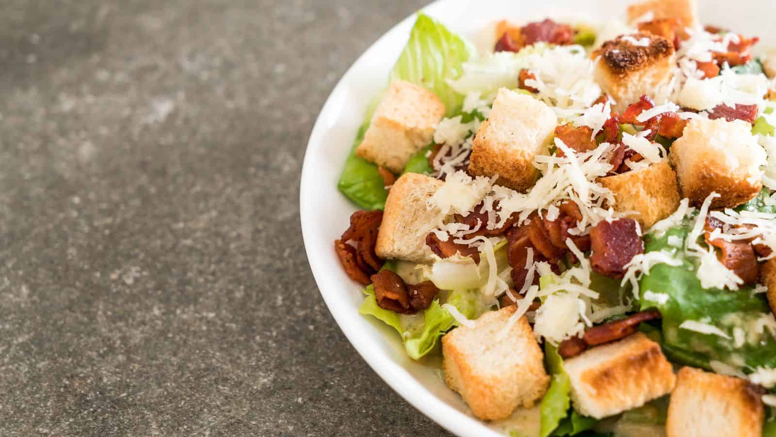 A Caesar salad in a white bowl on a gray surface, featuring romaine lettuce, croutons, grated cheese, and bacon pieces.