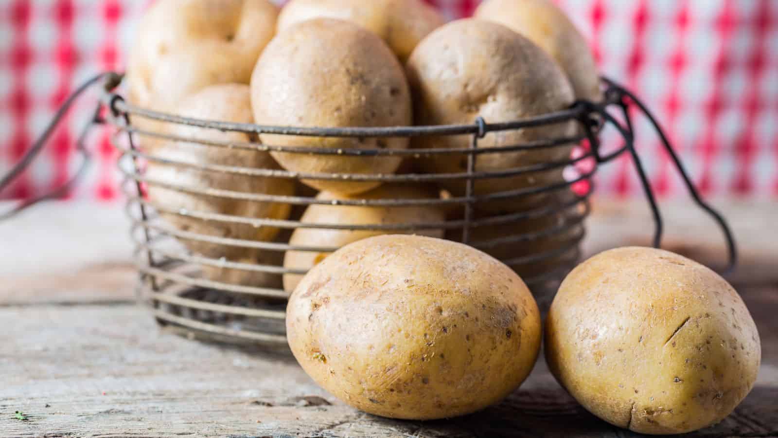 A wire basket filled with several brown potatoes sits on a wooden surface. Two potatoes rest outside the basket in the foreground. A red and white checkered pattern is visible in the blurred background.