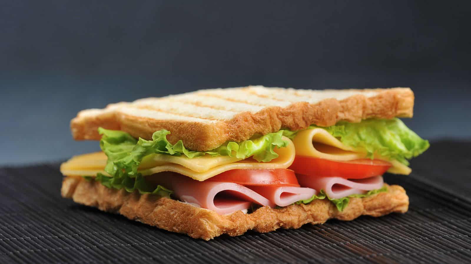 A sandwich with grilled toast, layered with lettuce, cheese slices, ham, and tomato slices. The sandwich is placed on a dark surface, showcasing its neatly stacked ingredients and lightly toasted bread.