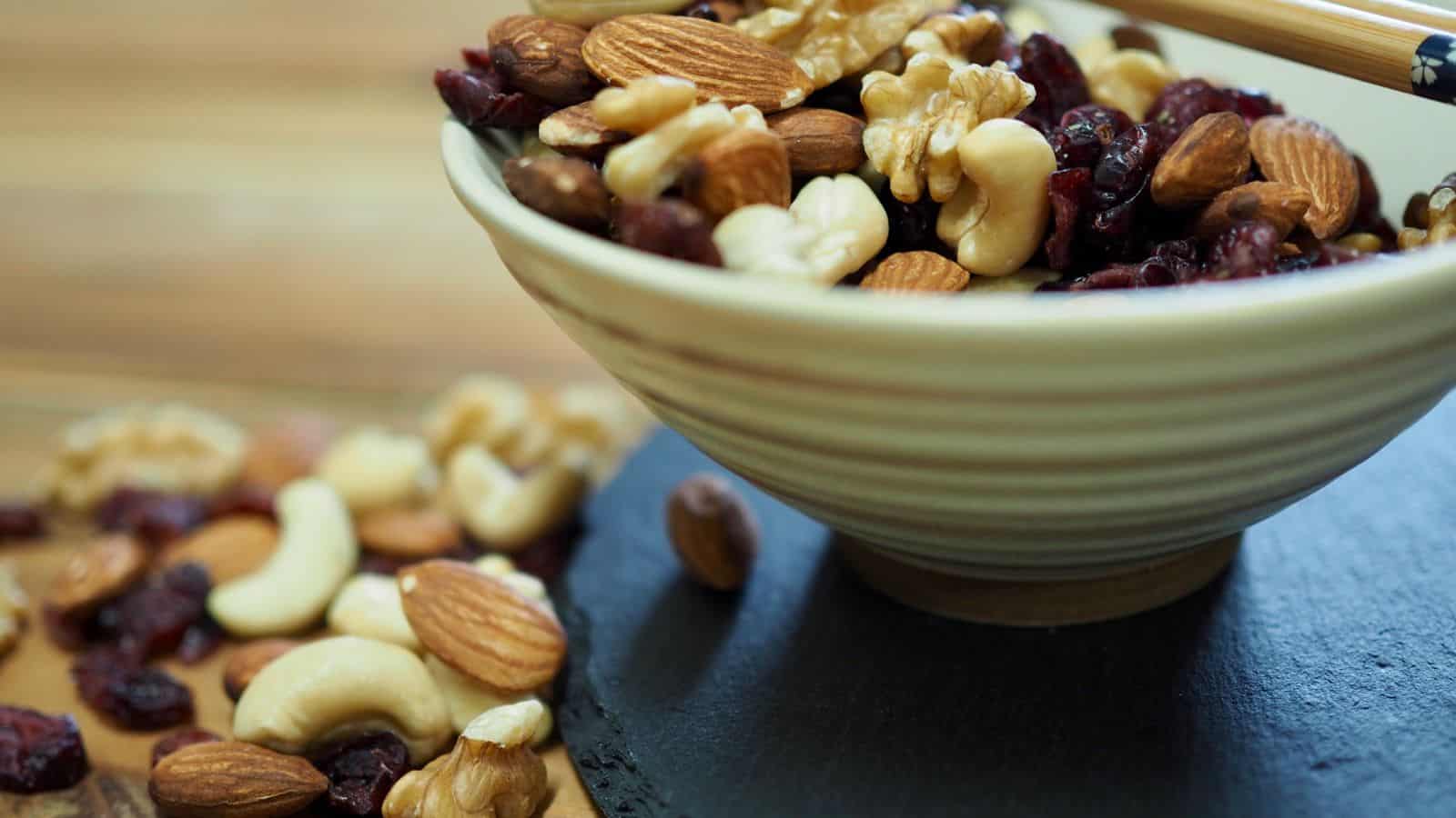 A ceramic bowl filled with a mixture of nuts and dried fruits, including walnuts, almonds, cashews, and dark raisins, sits on a dark surface. Additional nuts and dried fruits are scattered nearby on a wooden surface.