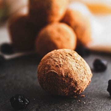 Close-up of a vegan chocolate truffle covered in cocoa powder on a dark surface. Other truffles are visible in the background, slightly out of focus. Small round dark items, possibly dried blueberries, are scattered nearby.