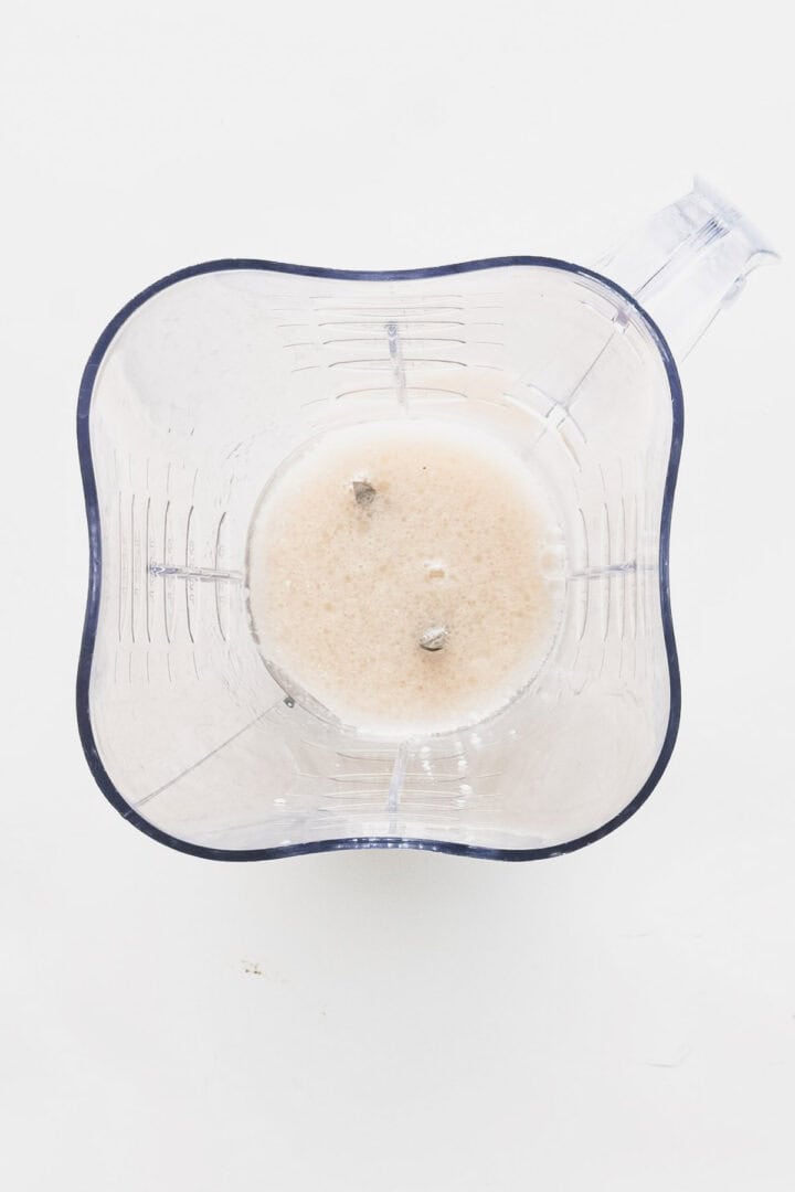 A transparent blender container viewed from above, partially filled with a light beige liquid containing small ice chunks and the essence of vegan chocolate truffles, rests on a pristine white surface.