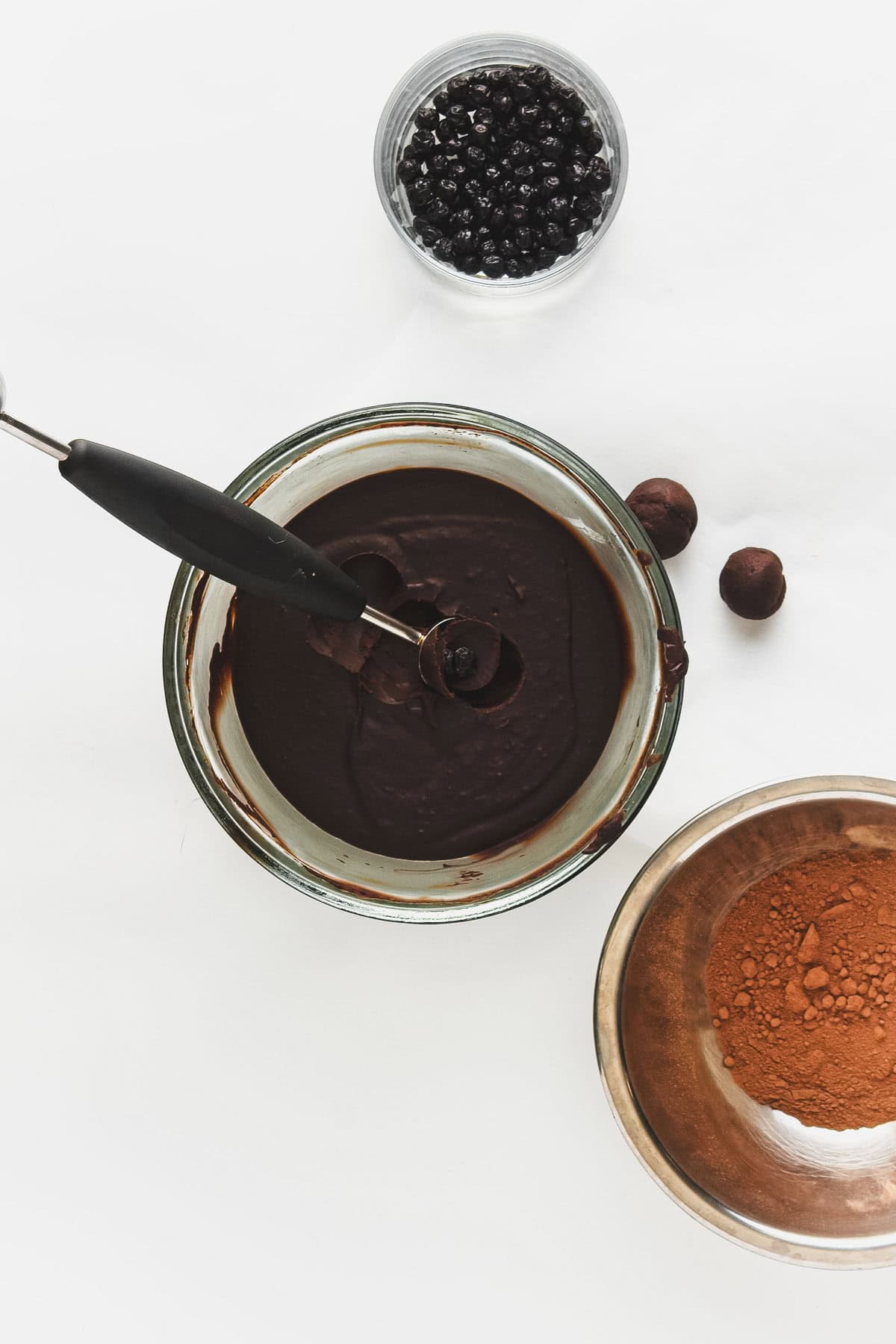 A glass bowl filled with chocolate ganache and a whisk awaits transformation into vegan chocolate truffles with blueberries. Nearby, a metal bowl brims with cocoa powder, whole nuts, and a small container of dark berries on a white surface.