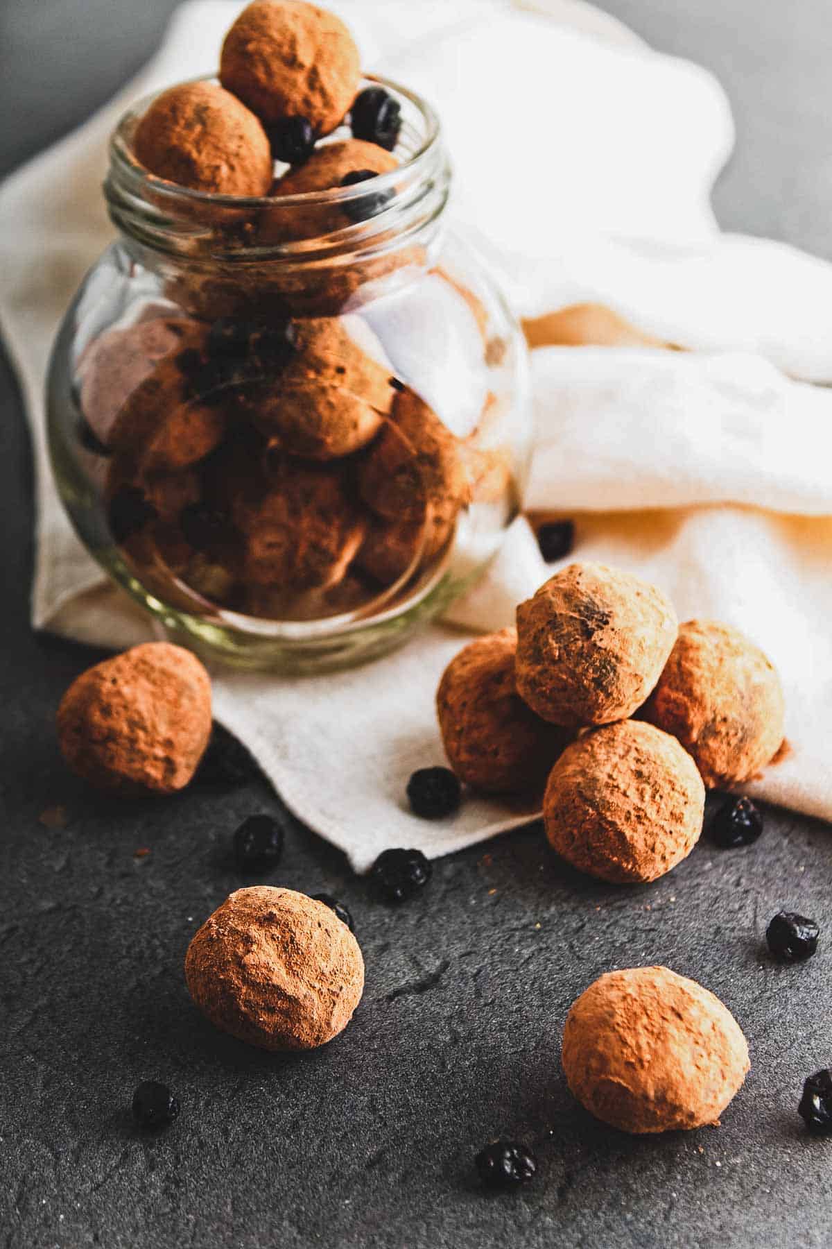 A glass jar filled with vegan chocolate truffles, dusted in rich cocoa, sits on a white cloth. A few truffles are scattered on a dark surface next to the jar, alongside small dried blueberries for an added burst of flavor.