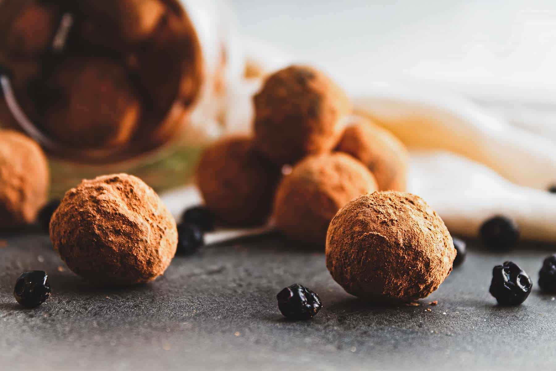 Vegan chocolate truffles are cocoa-dusted and scattered on a dark surface, mingling with small dried blueberries. A tipped container in the background suggests they've spilled out, with a soft, blurred cloth partially visible behind them.