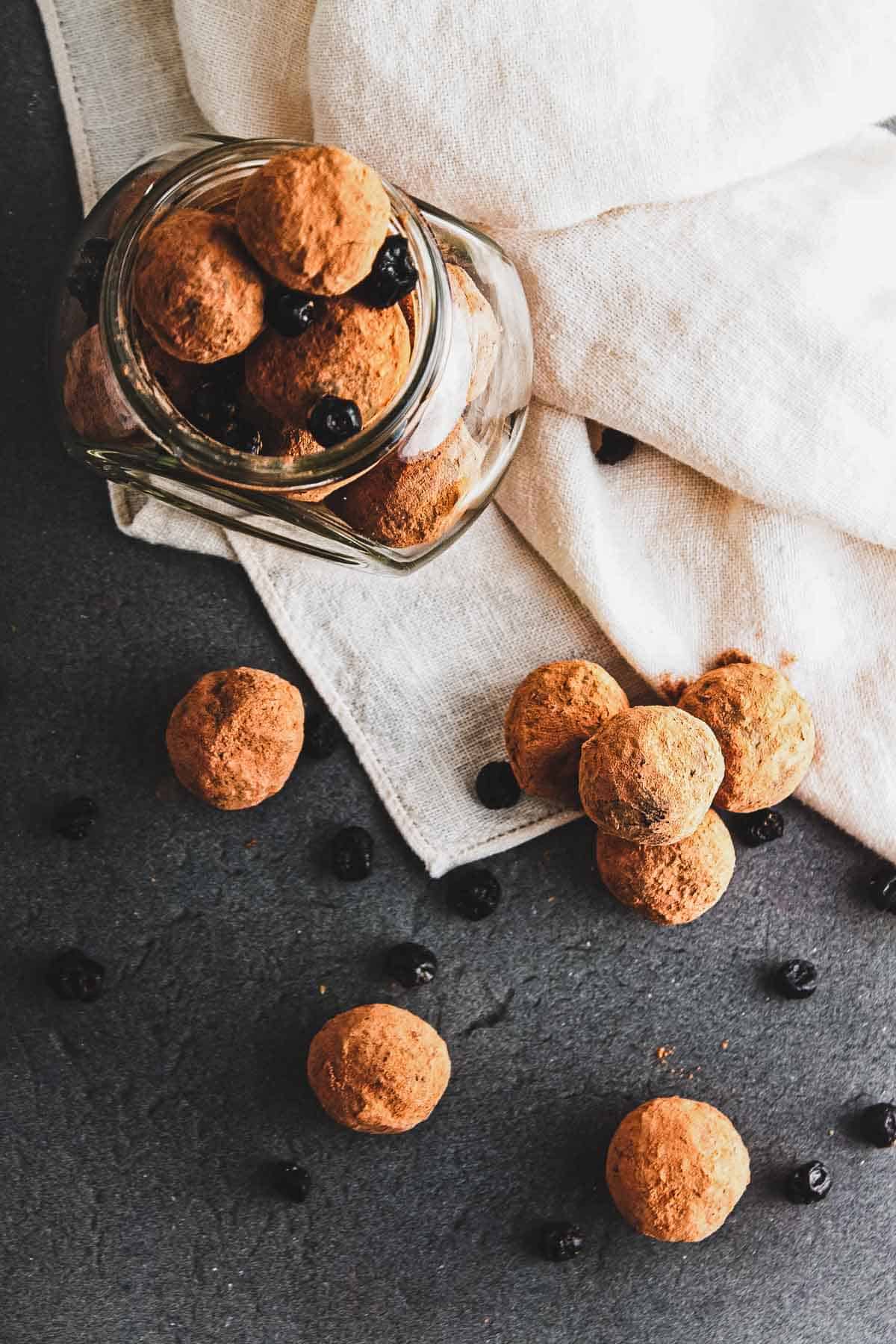 A glass jar tipped over on a dark surface, spilling vegan chocolate truffles dusted with cocoa and dried blueberries onto a white cloth. Extra truffles and blueberries are scattered around, creating a delightful mix of flavors.