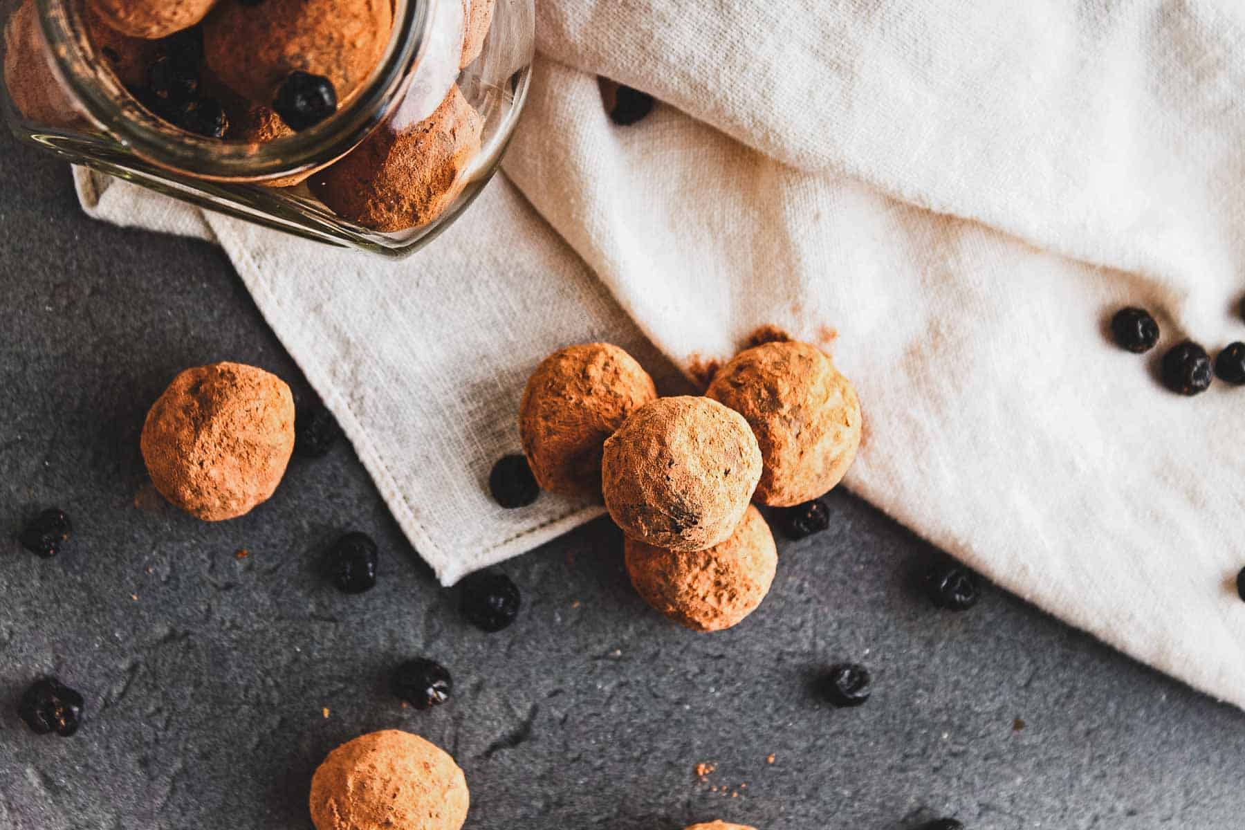 A group of round, brown vegan chocolate truffles is scattered on a dark surface, with some partially inside a jar. A white cloth lies nearby, and small dark blueberries are scattered around the truffles.