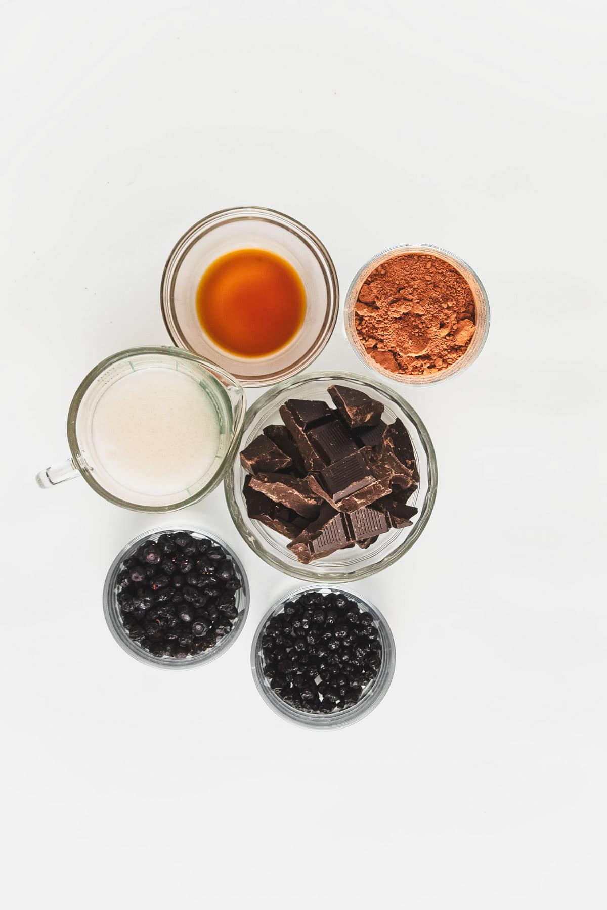 A variety of ingredients are arranged on a white surface, including a bowl of sugar, cocoa powder, chocolate chunks for vegan chocolate truffles with blueberries, and bowls of fresh blackberries and blueberries next to an amber-colored liquid.