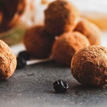 Chocolate truffles coated with cocoa powder are placed on a gray surface. Some of the truffles and small dried berries are in the background, slightly out of focus. A light fabric is draped behind them.