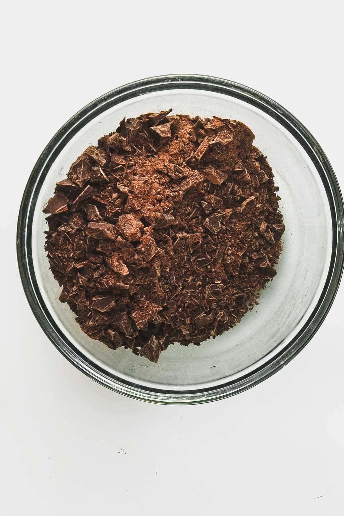 A clear glass bowl filled with chopped vegan chocolate pieces, set against a plain white background, hints at the creation of indulgent truffles with blueberries.