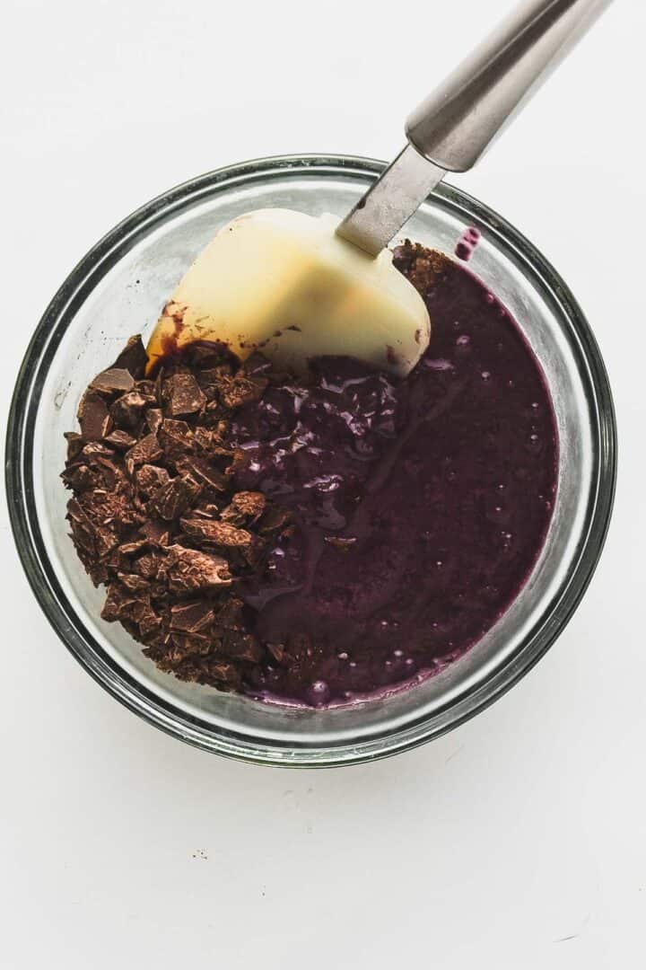 A glass bowl holds a purple batter for vegan chocolate truffles, partially mixed with chopped chocolate and blueberries on one side. A white spatula with a metal handle rests inside the bowl, surrounded by delicious ingredients.