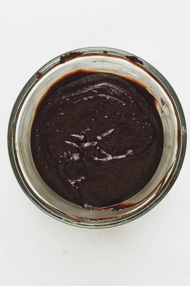 Glass bowl containing thick, dark brown vegan chocolate truffle batter with hints of blueberries on a white background. The surface is smooth yet displays enticing textures, promising a delightful treat.