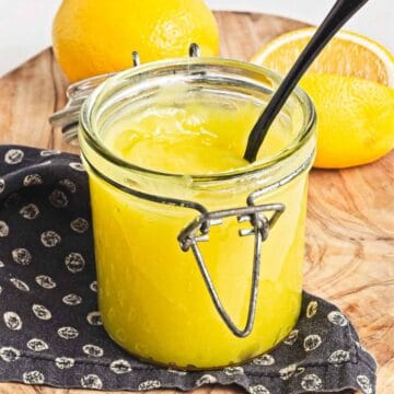 A glass jar filled with vibrant vegan lemon curd sits on a wooden surface, a black spoon resting inside. Behind it, a whole lemon and a half-cut lemon add to the citrus display. The jar stands proudly on a dark cloth adorned with small lemon patterns.