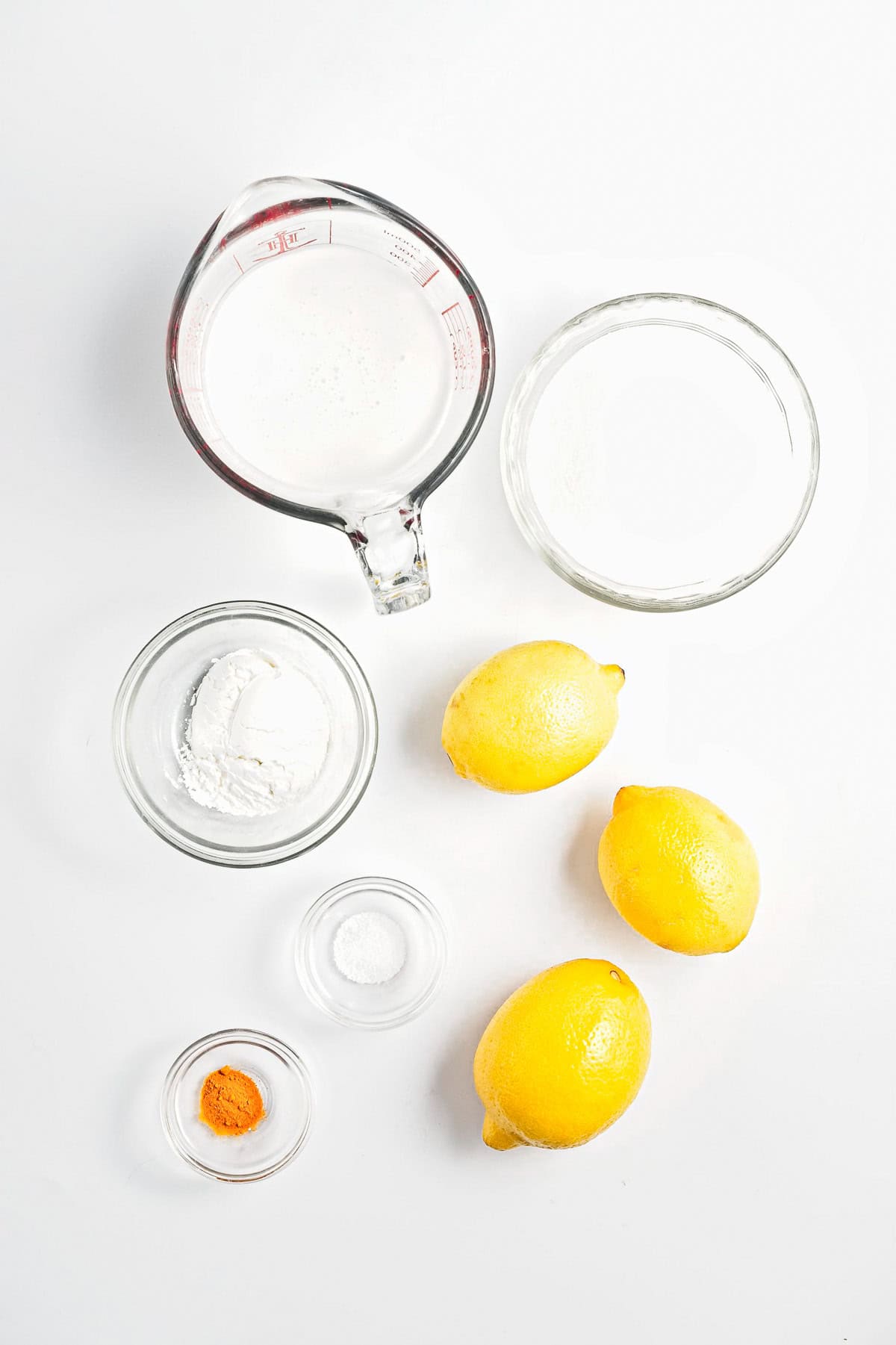 Top-down view of ingredients on a white surface: a glass measuring cup with liquid, a clear bowl with sugar, three lemons for lemon curd, a bowl with white powder, a small bowl of salt, and a tiny bowl featuring vegan orange powder.