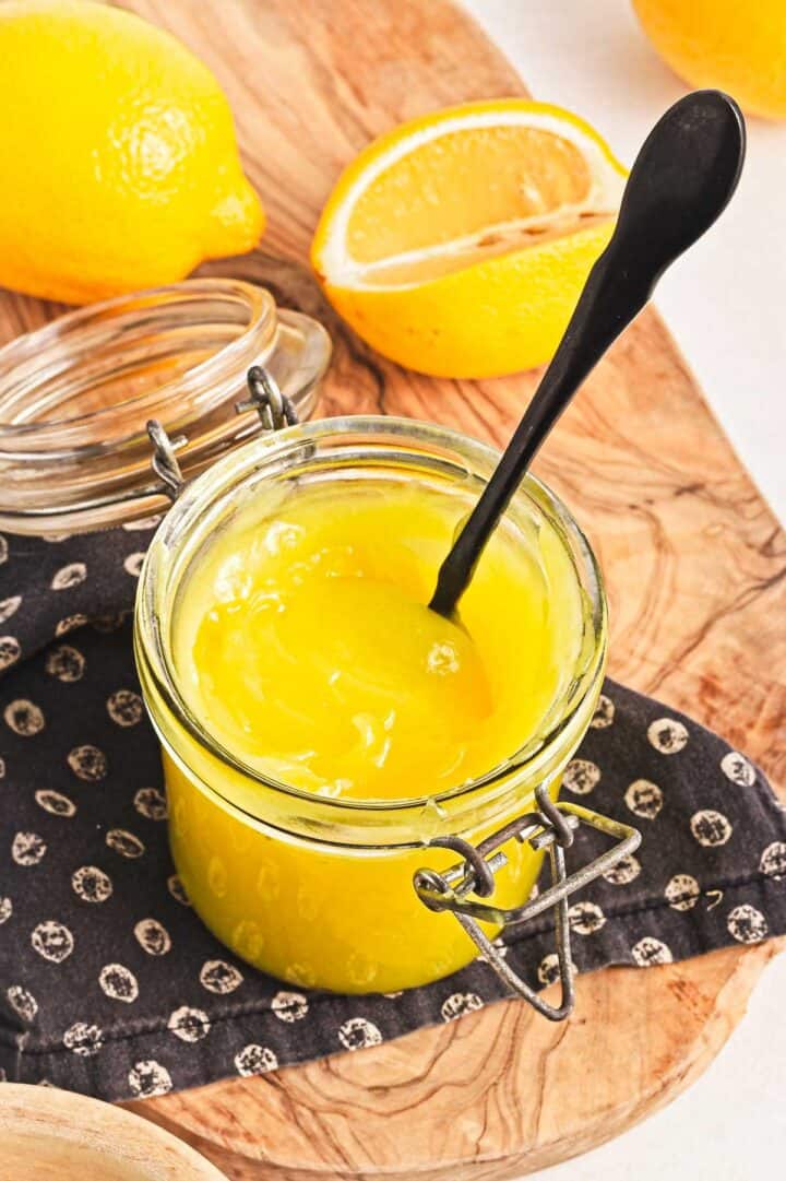 A jar of vibrant yellow vegan lemon curd with a black spoon rests on a patterned cloth on a wooden board. Behind it, a whole lemon and a halved lemon add to the scene. The jar's hinged lid is slightly open against the light background.