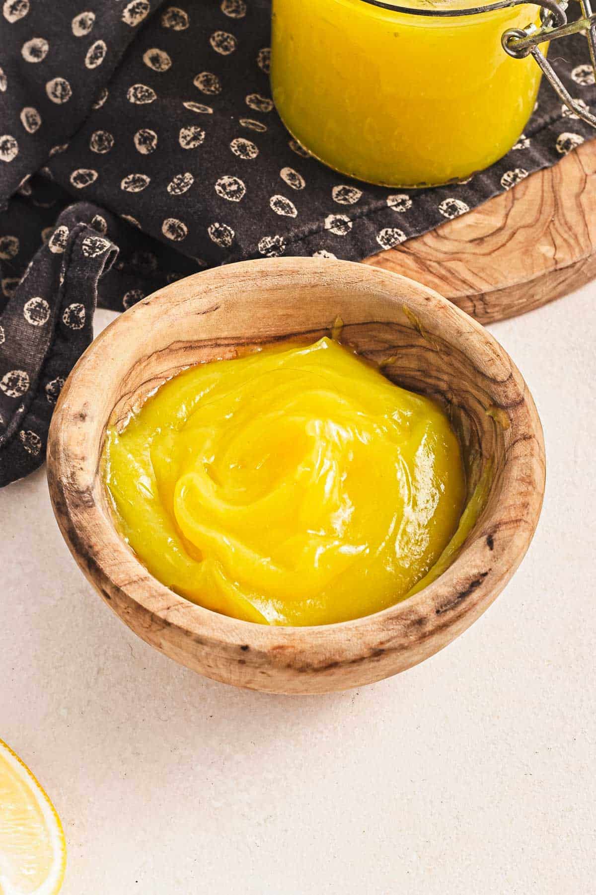 A wooden bowl brimming with silky, yellow lemon curd rests on a light surface. In the background, a piece of fabric adorned with lemon patterns and a jar of vegan lemon curd are visible. A lemon slice peeks in from the left, adding a zesty touch.