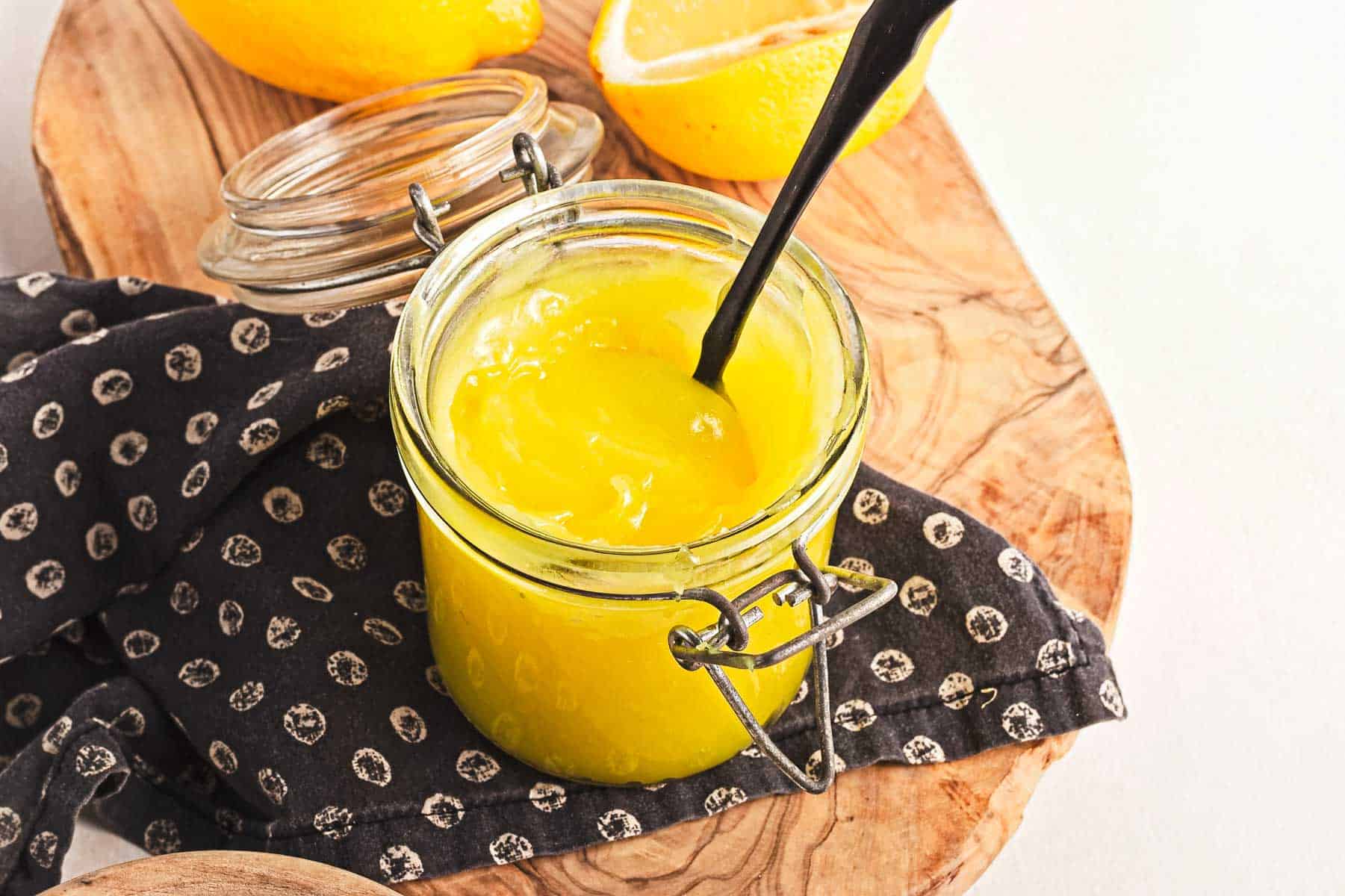 A jar of bright yellow lemon curd with a black spoon inside sits on a wooden board. A patterned cloth is underneath the jar, and a halved lemon and another whole one add zest to the scene. The open lid rests nearby, inviting you to indulge in this tantalizing vegan treat.