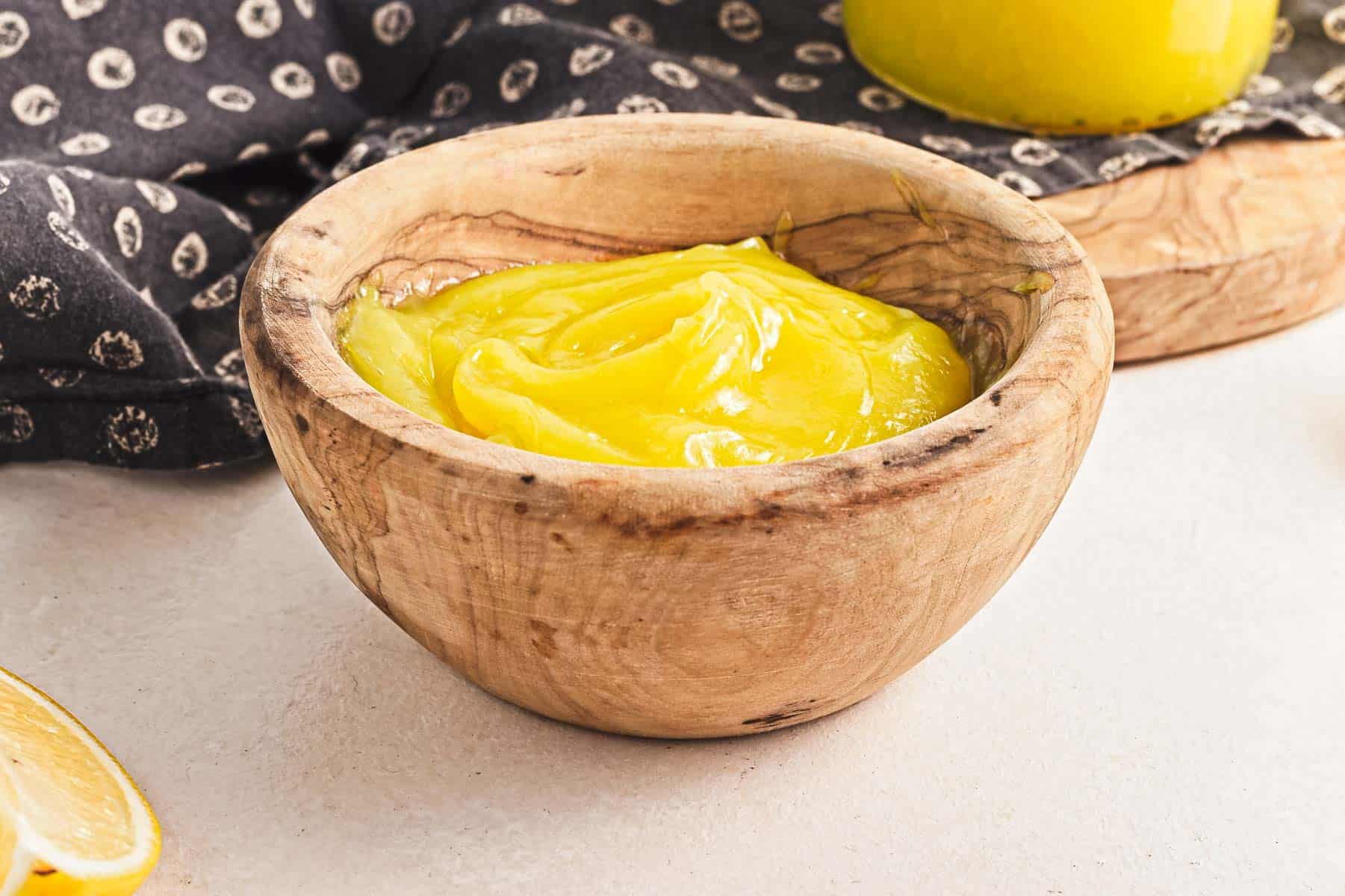 A wooden bowl brimming with vegan lemon curd sits on a light surface. A slice of lemon and a patterned dark cloth add charm to the scene, while another jar of the vibrant yellow treat peeks into view.