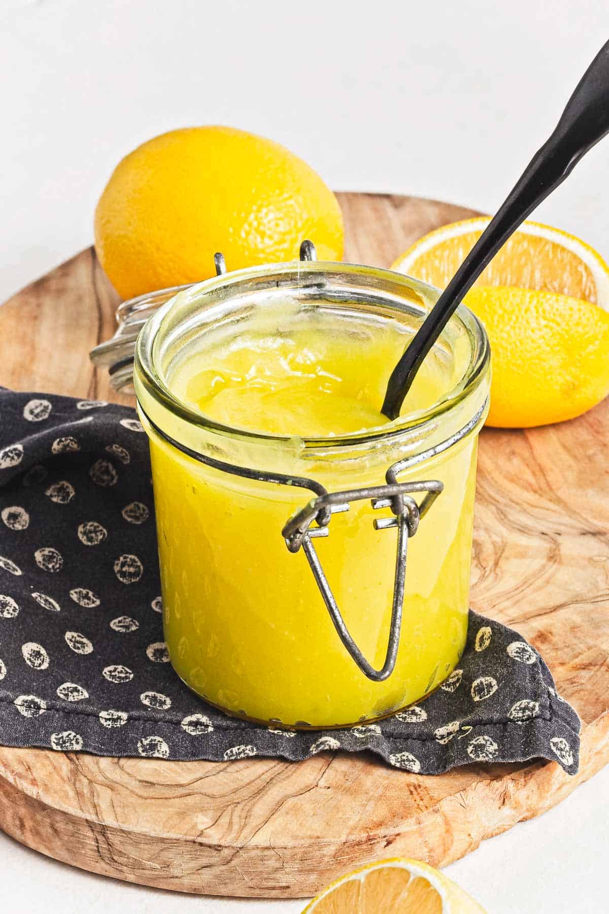 A glass jar brimming with vegan lemon curd sits on a wooden board. A black spoon rests inside, and a dark patterned cloth lies beneath. Whole and sliced lemons are artfully arranged in the background, adding a fresh touch to the scene.