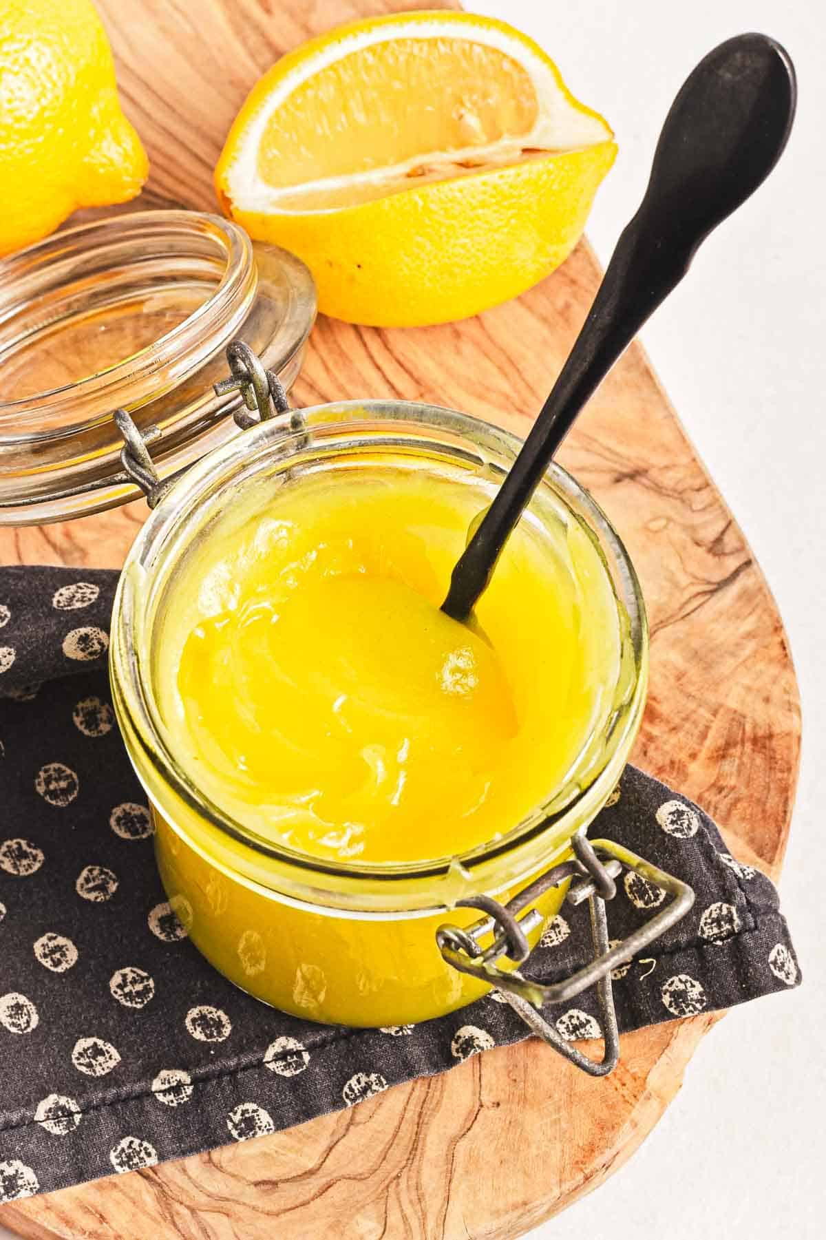 A jar of vegan lemon curd with a black spoon rests on a wooden board, complemented by a black-and-white patterned cloth beneath it. A halved lemon sits in the background.