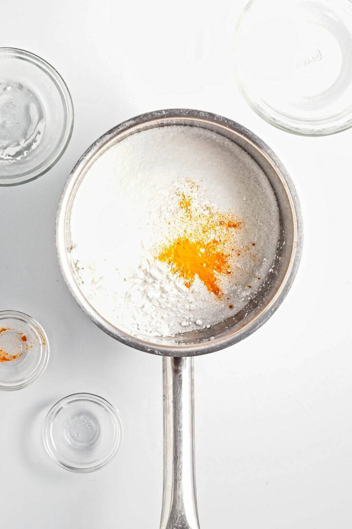 A metal saucepan contains white powder with a hint of yellow-orange powder, reminiscent of lemon curd. Surrounding the saucepan are empty glass bowls, all resting on a pristine white surface.