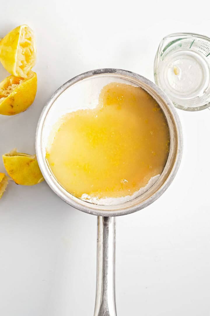 A saucepan filled with melted butter and lemon zest sits on a white surface, hinting at the beginning of a delicious lemon curd. To the left lie squeezed lemon halves, while to the right, a small glass container holds what seems to be cream or buttermilk.