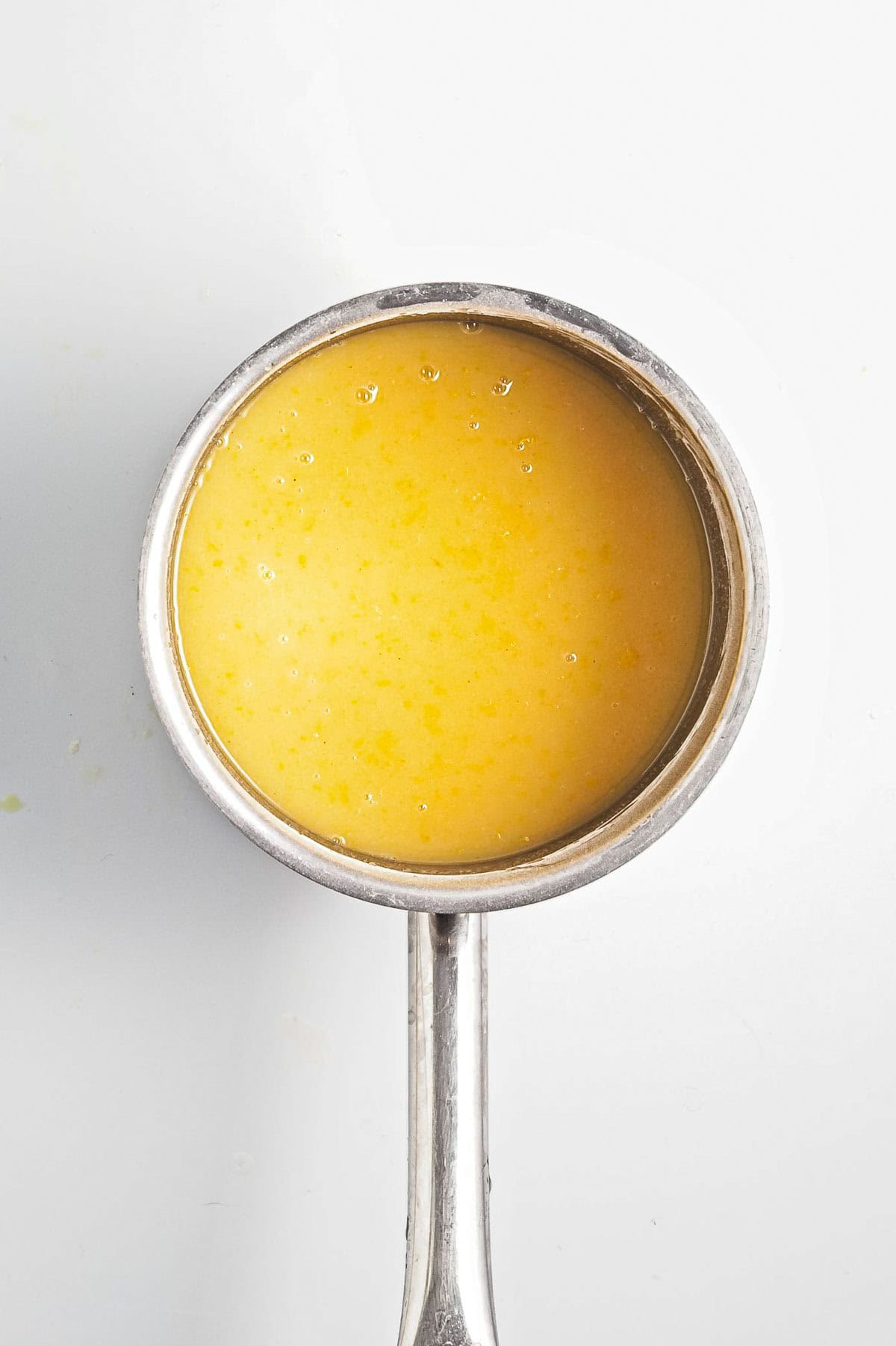 A metal ladle filled with a vibrant yellow liquid, possibly vegan broth or sauce with a hint of lemon, rests on a white surface. The liquid displays a slightly textured appearance, with small air bubbles visible on the surface.