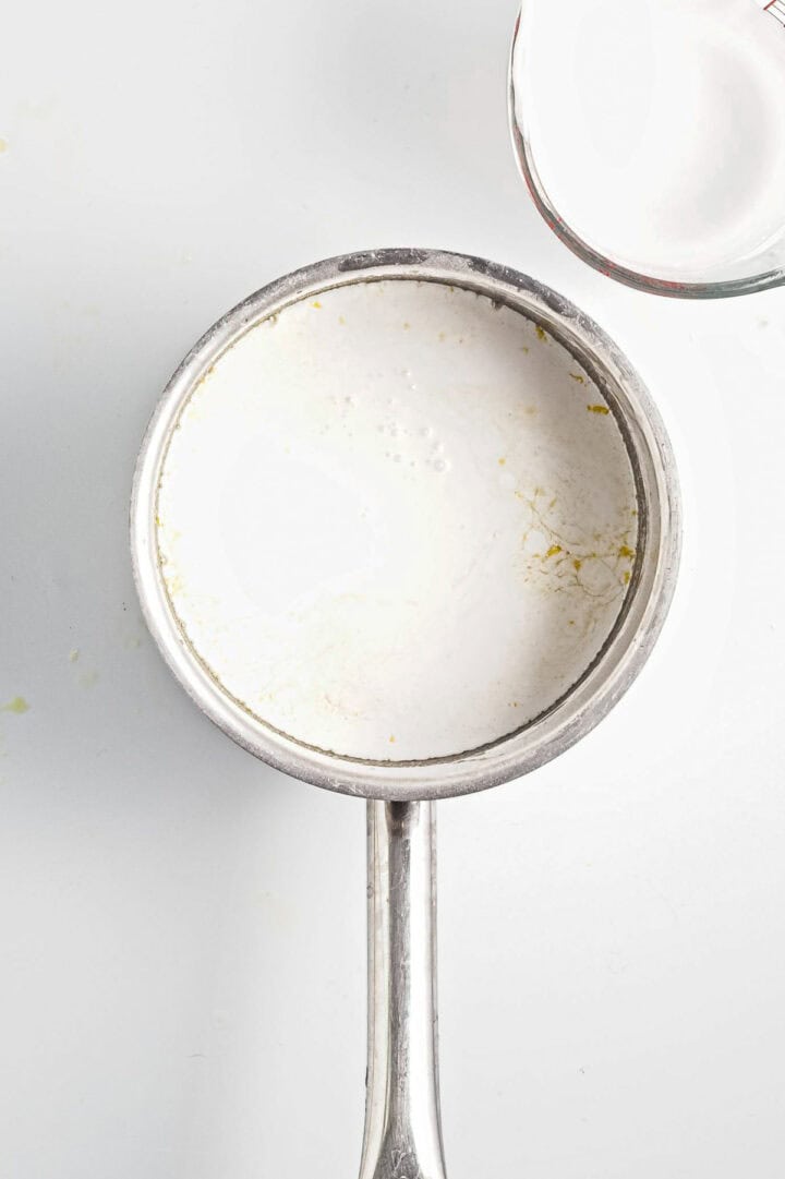 A small steel saucepan containing white liquid sits on a white surface, hinting at the comfort of a homemade vegan treat. The liquid shows slight yellowish stains around the edges, reminiscent of lemon curd. Part of a clear glass container with similar contents peeks from the top right corner.