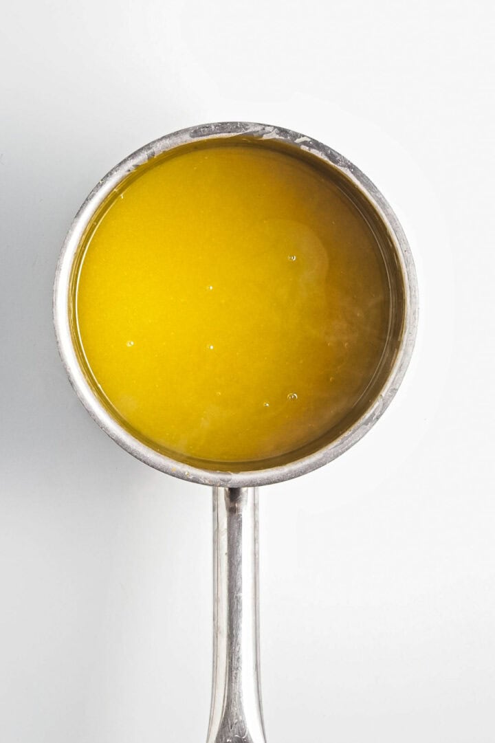 A top-down view of a metal saucepan filled with a vibrant yellow lemon curd against a plain white background, showcasing its smooth vegan texture.