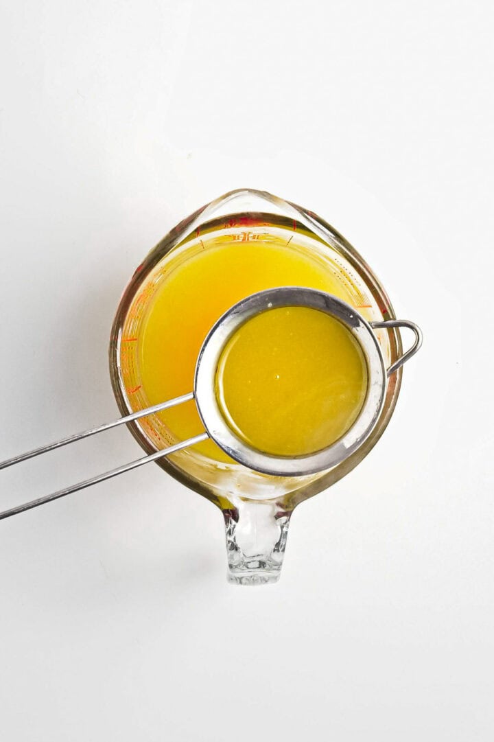 A clear measuring cup brims with vibrant orange-yellow lemon curd. A metal strainer rests atop, capturing the essence of a smooth vegan filtering process. The plain white background complements the scene’s simplicity and purity.