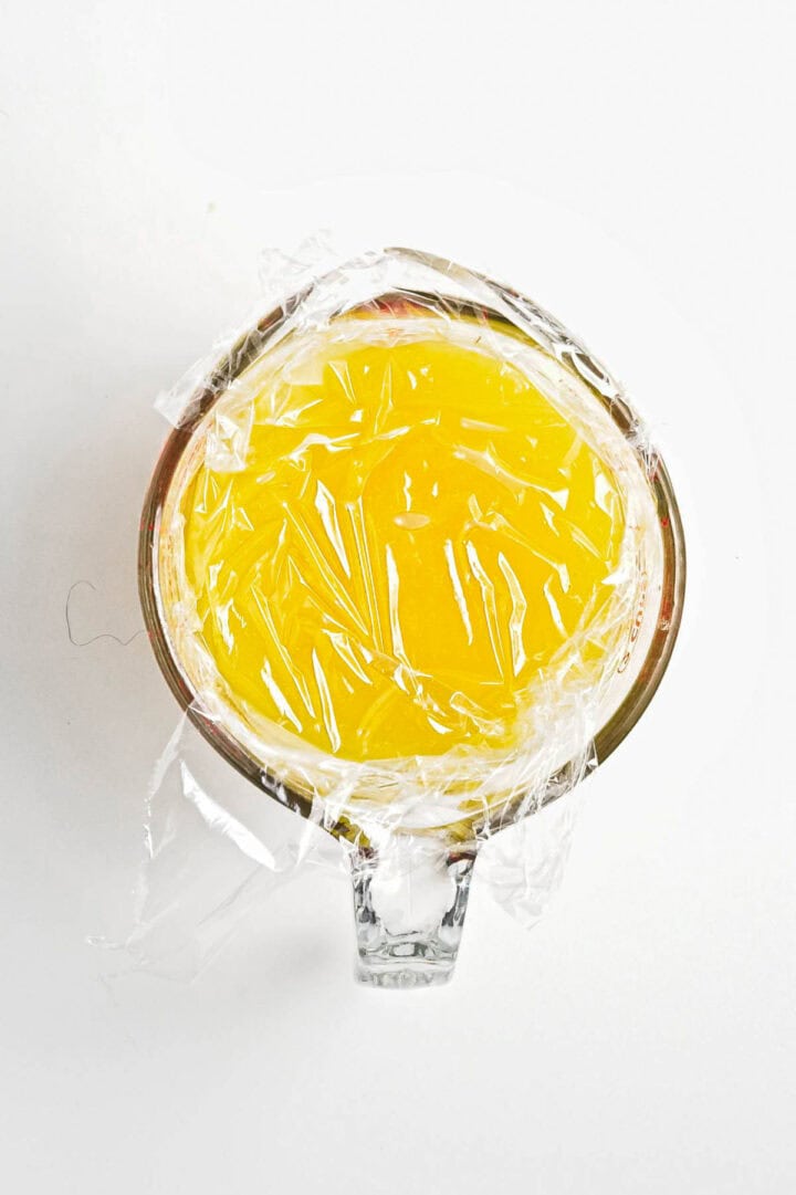 A glass measuring cup filled with bright yellow liquid, possibly a tangy lemon juice or vegan broth, is sealed with clear plastic wrap. The cup rests on a pristine white background.