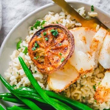 A plate featuring grilled scallops on skewers atop brown rice, garnished with a slice of charred lemon. For those hooked for life on seafood recipes, green beans sit alongside. A fork rests nearby, and a small bowl of salt is close at hand on a linen cloth.