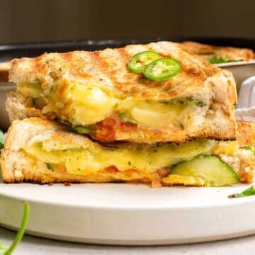 A grilled cheese sandwich with visible slices of jalapeños, cucumber, and tomatoes is cut in half and stacked on a white plate. A pan and a kitchen towel are in the background, and a few loose jalapeños and cilantro leaves are scattered around.