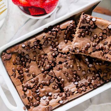 A rectangular baking dish filled with chocolate brownies topped with chocolate chips. One brownie is partially lifted out. A bowl of sliced strawberries and two forks are beside the dish on a white tablecloth.