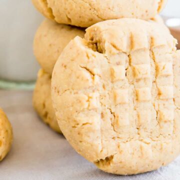 A stack of three classic peanut butter cookies with a crisscross pattern evokes a nostalgic 1970s charm. The top cookie has a bite taken out of it, and a glass of milk lingers in the background on the white surface and light blue cloth beneath.