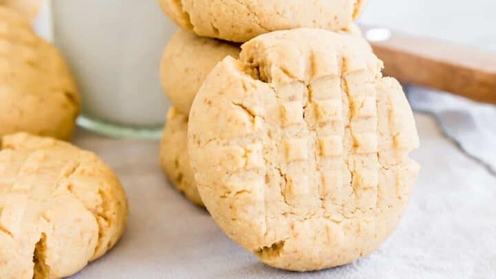 A stack of three classic peanut butter cookies with a crisscross pattern evokes a nostalgic 1970s charm. The top cookie has a bite taken out of it, and a glass of milk lingers in the background on the white surface and light blue cloth beneath.