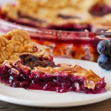 A slice of grape pie is on a white plate, set on a wooden cutting board. The pie has a golden, flaky crust with a purple grape filling. A bunch of grapes is beside the plate, and the rest of the pie is visible in the background.