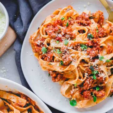 A plate of fettuccine pasta topped with a red meat sauce and garnished with grated cheese and herbs. A bowl of grated cheese and another dish of pasta are visible on a wooden surface alongside a dark cloth.