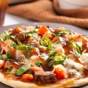 A close-up of a circular pizza topped with diced tomatoes, chopped cilantro, pieces of meat, and drizzled with sauce. It sits on a dark surface with a brown cloth and a beverage in the background.