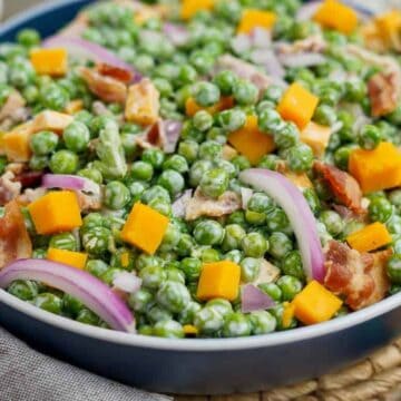 A bowl filled with a colorful salad, featuring green peas, diced yellow cheese, red onion slices, and crispy bacon pieces. The dish is set on a woven placemat with a small dish in the background.