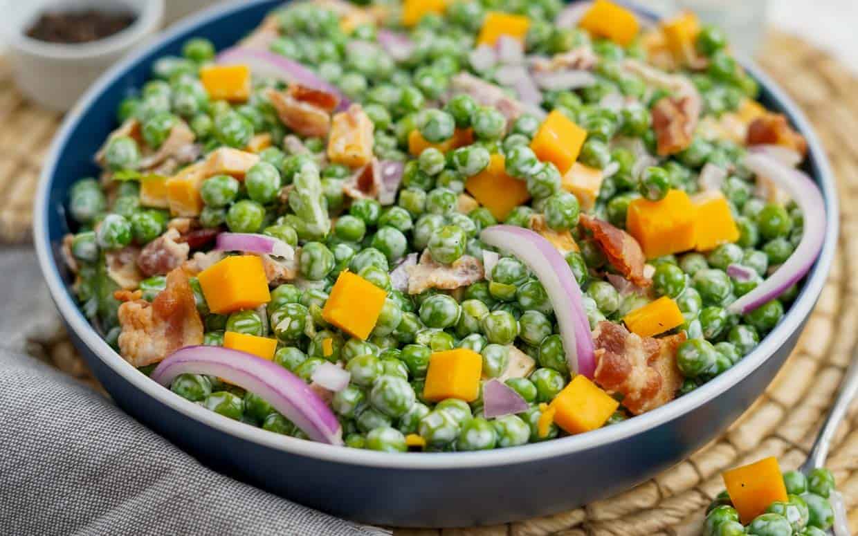 A bowl filled with a colorful salad, featuring green peas, diced yellow cheese, red onion slices, and crispy bacon pieces. The dish is set on a woven placemat with a small dish in the background.