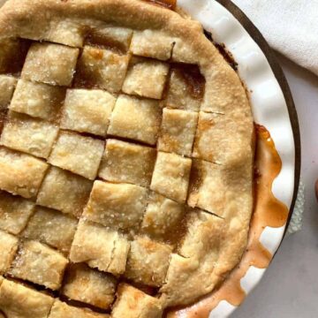 A freshly baked peach pie with a golden lattice crust sits in a white pie dish. Two whole peaches and a small dark bowl with green leaves are on the marble surface nearby. A folded white napkin is partially visible in the top right corner.