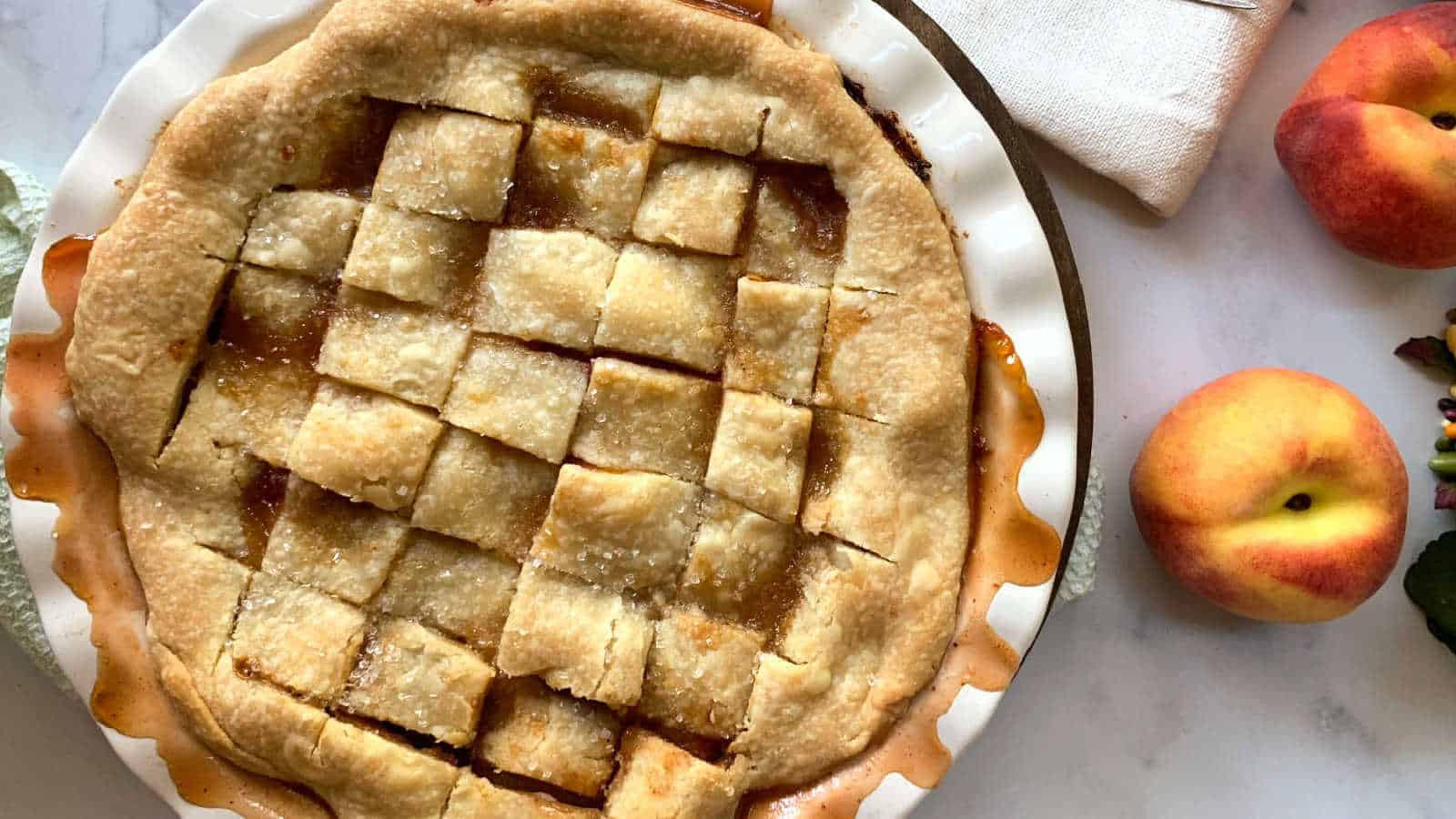 A freshly baked peach pie with a golden lattice crust sits in a white pie dish. Two whole peaches and a small dark bowl with green leaves are on the marble surface nearby. A folded white napkin is partially visible in the top right corner.